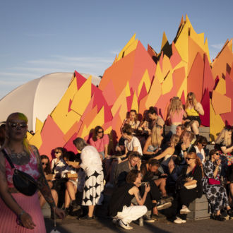 Dozens of people sit on stacks of boxes in front of a colourful artwork.