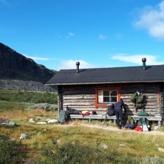 Several people with hiking gear are in front of a wooden building with mountains in the background.