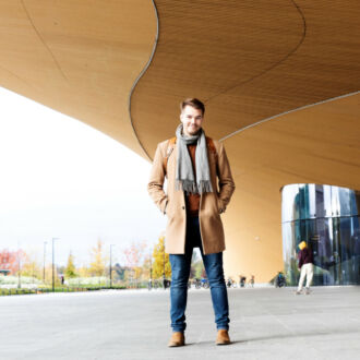 A man in a coat and scarf stands in front of a building made of wood and glass.